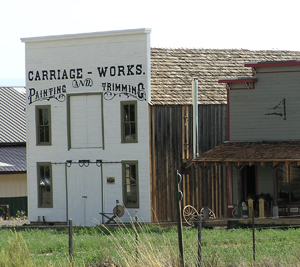 A view of the building with white square facade with two floors, doors in the center and windows on the side of both and above a sign in the rear is a wood walled structure with gable roof.