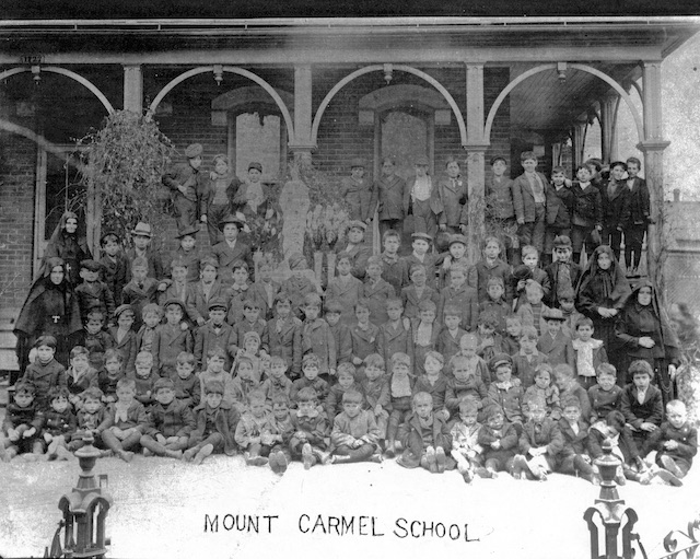 Historic photograph of the students and missionary sisters at the Mount Carmel School around 1902. There is a large group of children of all ages sitting on the ground in front of the school, while older children stand on the front porch alongside sisters of the Cabrini missionary school.