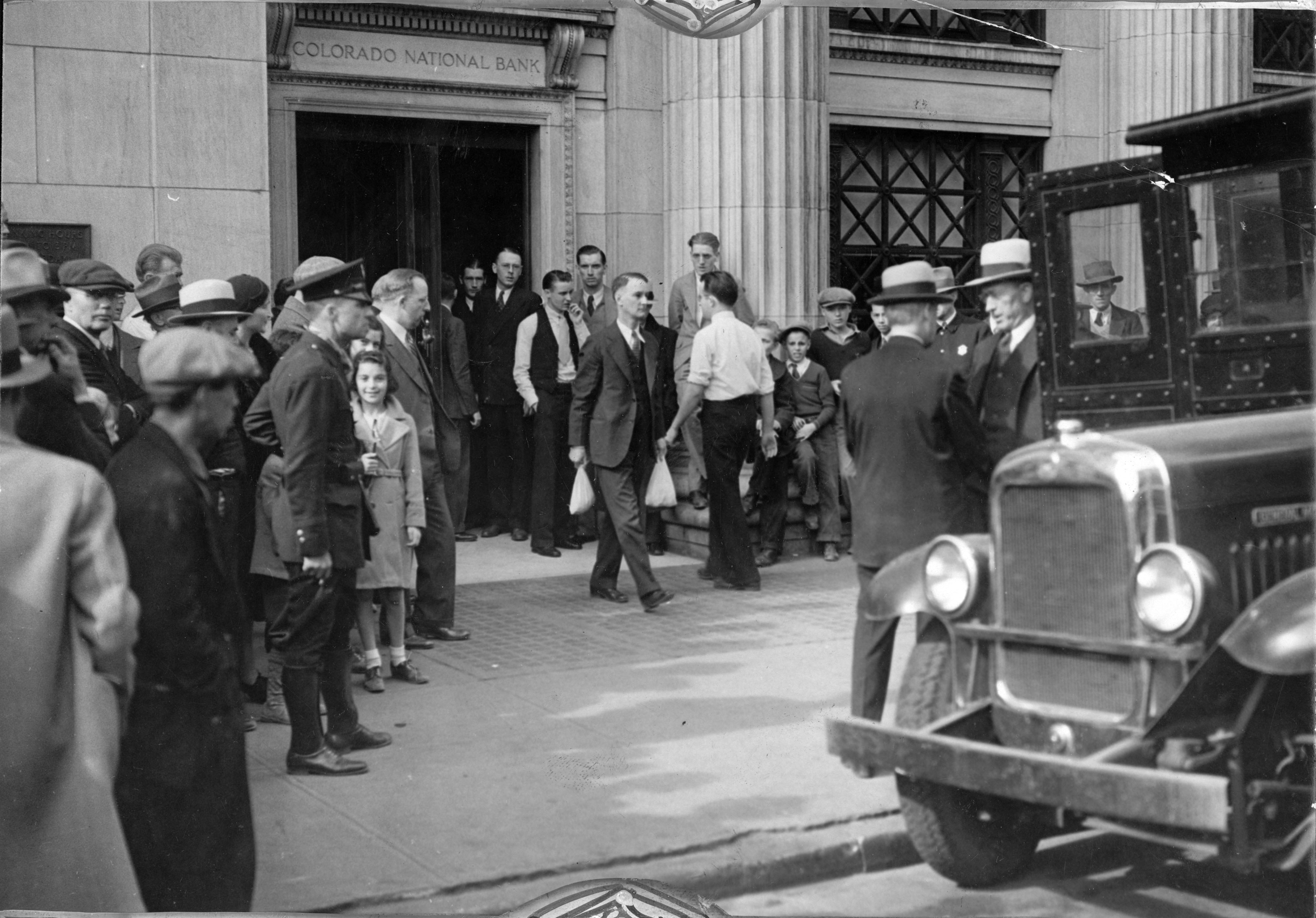 A photo taken in 1933 of the Colorado National Bank. The shot was taken shortly after the bank reopened from a bank holiday, and many people of all ages stand outside of the bank and appear to be waiting to enter the bank, or are walking past along the sidewalk. Most of those pictured are dressed in coats, suits, and hats. The front end of a car is in the foreground of the photo.