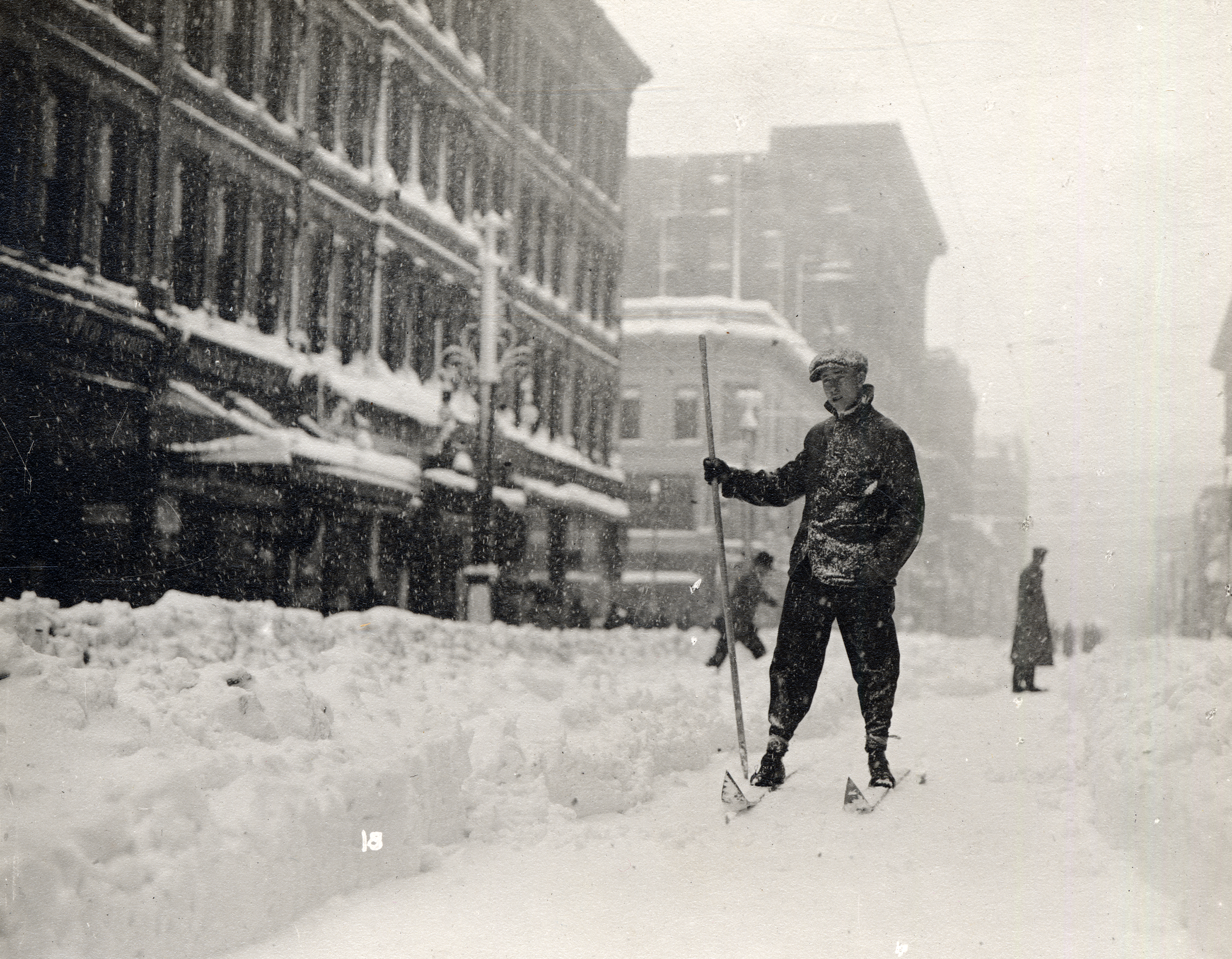 Photo of a young man on skis and holding a pole while skiing through the snow-covered street of downtown Denver. Snow is falling, and there is a narrow path cleared between large snowdrifts on what would be the city street.