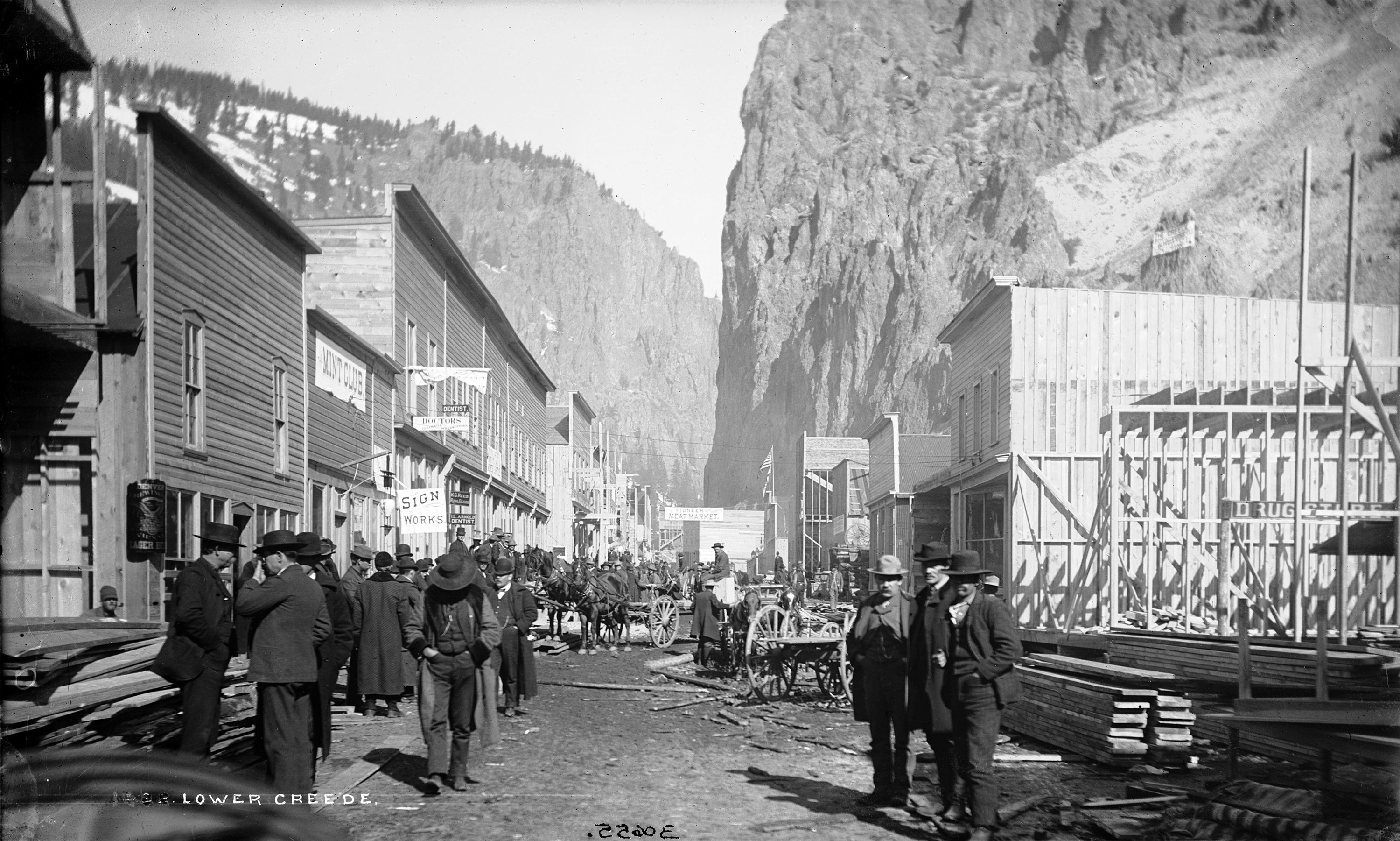 Photo of a street in downtown Creede, Colorado, in the 1890s. Several wooden buildings line each side of the dirt street, while other buildings are under construction and pallets of building materials sit on the sides of the street. Men dressed in suits and hats are seen walking and standing around talking with others as horse-drawn wagons traverse the road.