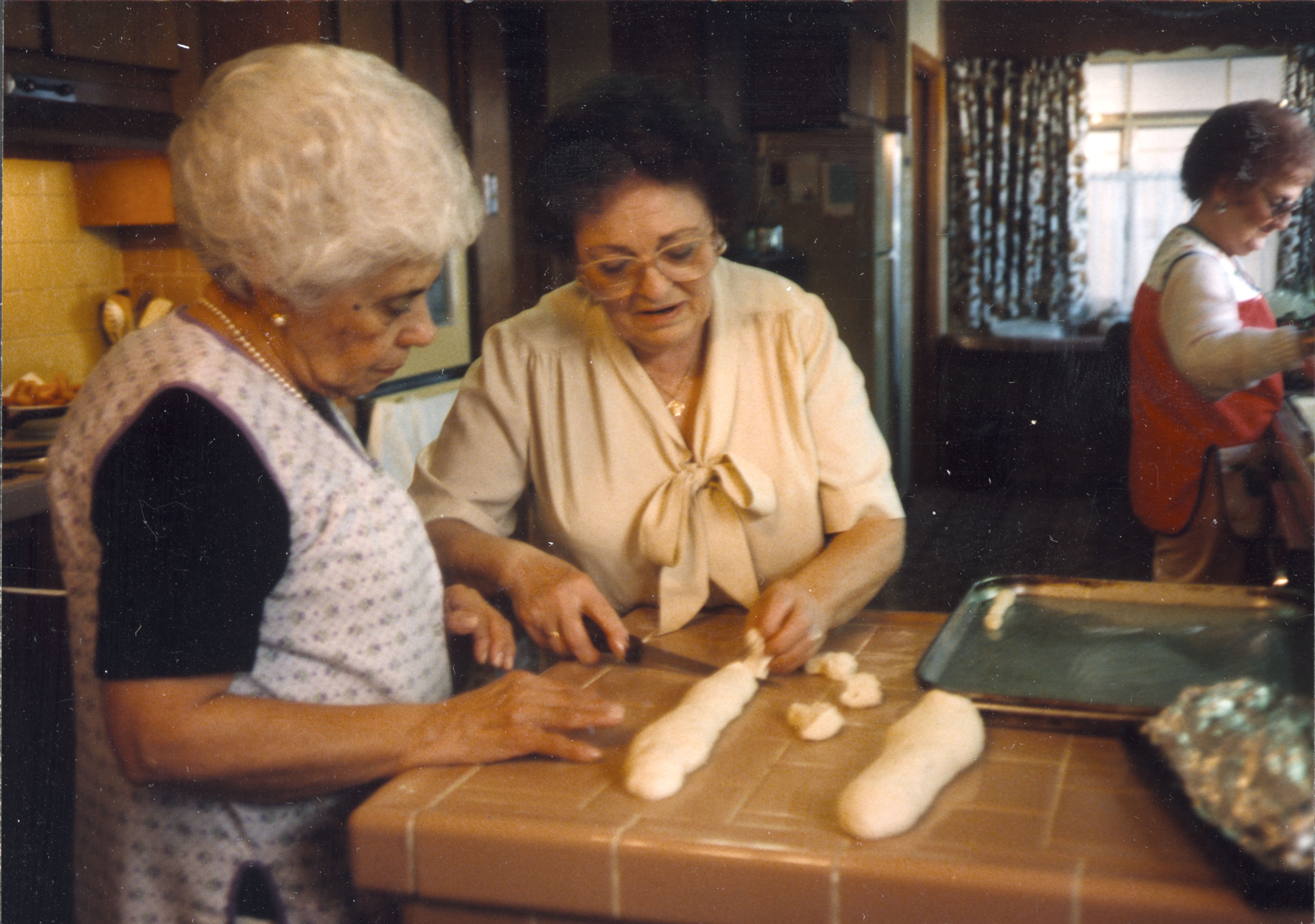 Photo of three women, Edna Canino, Mary Rotola, and Carmella Canino Preparing Bread for St. Joseph Table. The women are wearing aprons and working together at a tiled kitchen counter to shape dough into bread loaves.