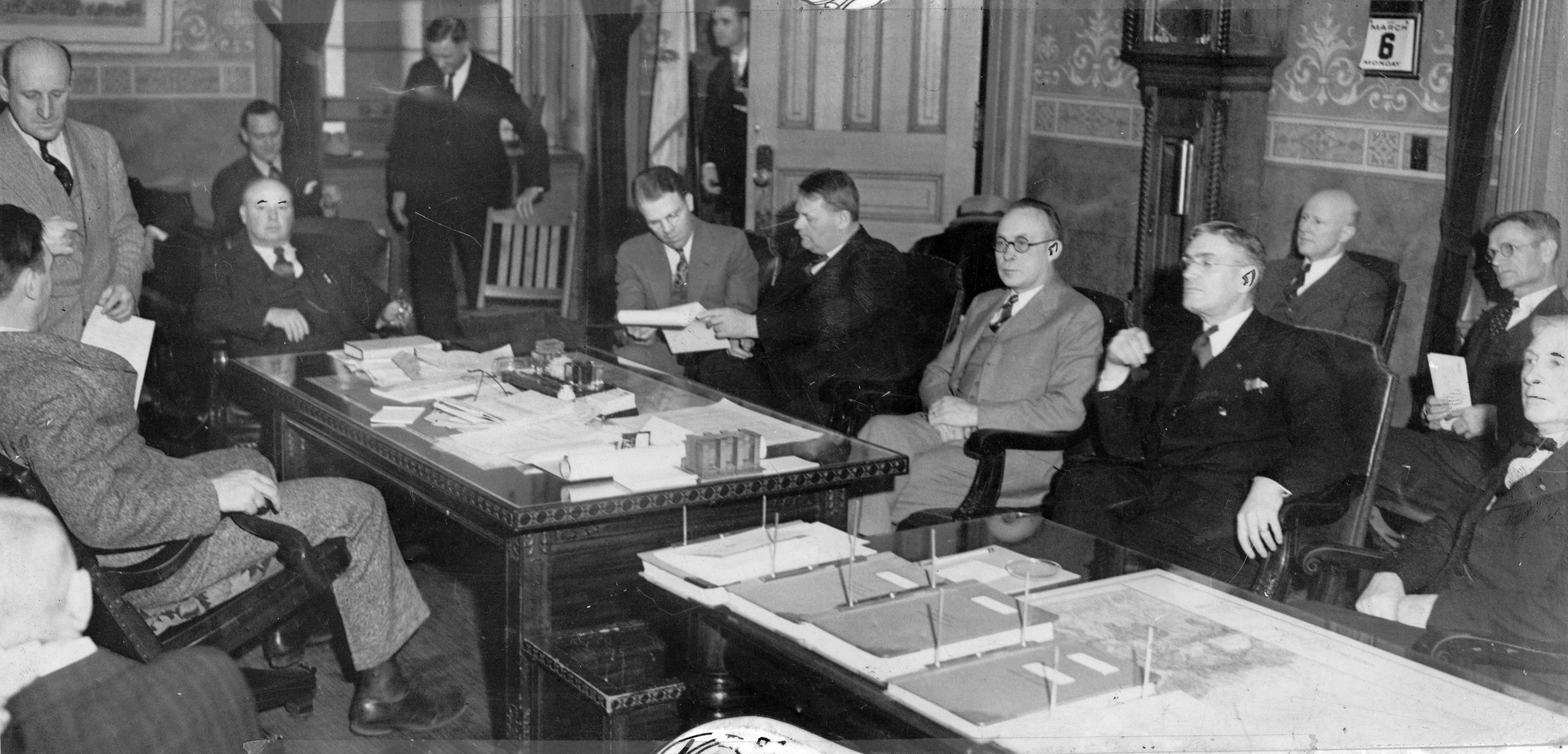 A photograph of the office of Governor Edwin C. Johnson during a meeting with Colorado bankers. There are at least 13 men present, sitting in chairs around a desk and adjacent table. The Governor's desk is scattered with papers, while neat stacks of paper and notebooks are laid out on the table next to it. The men are all dressed in suits, and some appear to be listening to someone speak, while a few men talk among themselves. 