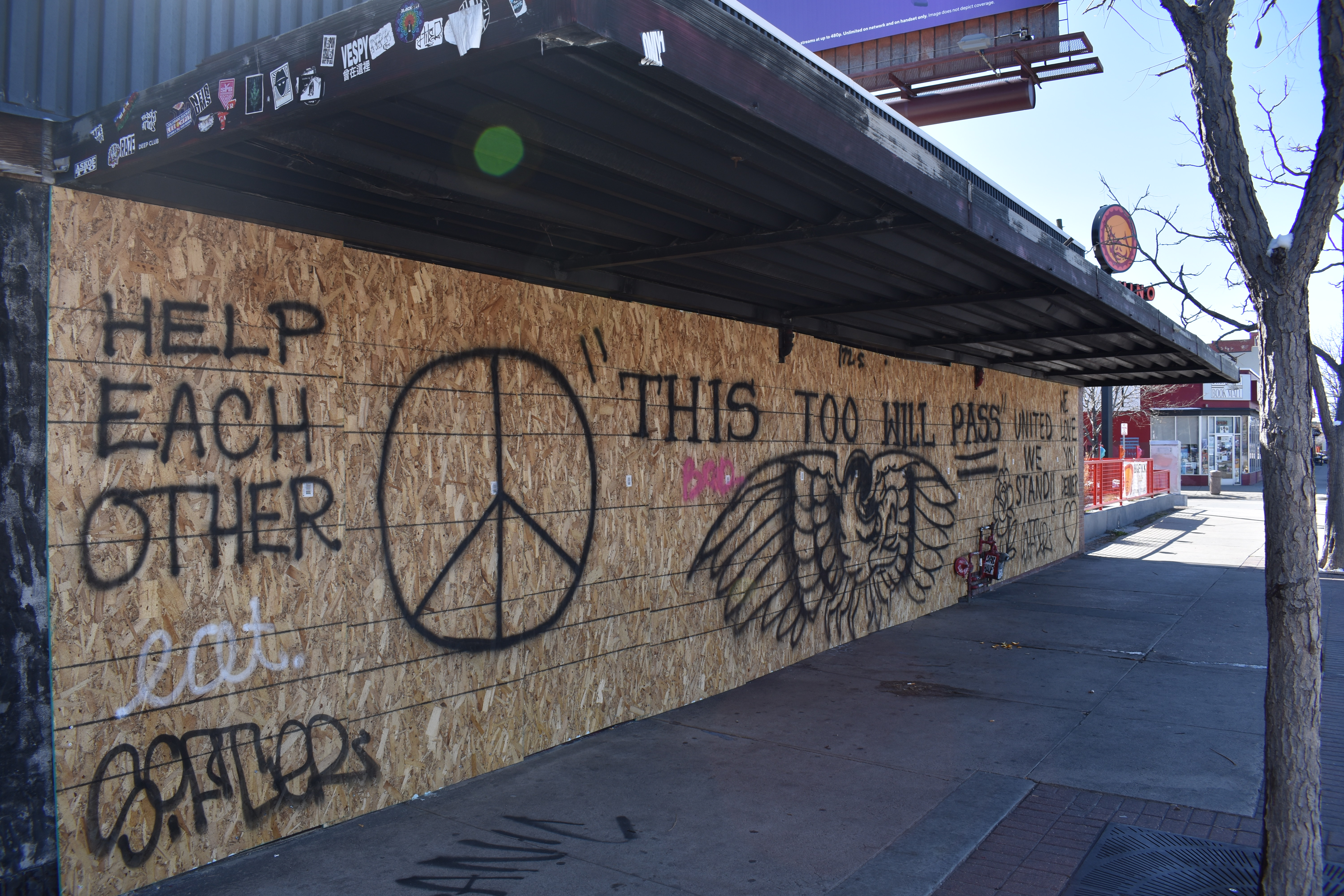 Photo of Think Tank Tattoo, with windows boarded up due to closure during the 2020 pandemic. The main image spray painted on the boards is an eagle and "THIS TOO WILL PASS". Right side detail consists of black outline of a flower, heart and the words, "UNITED WE STAND" and "WE LOVE YOU DENVER!!" 