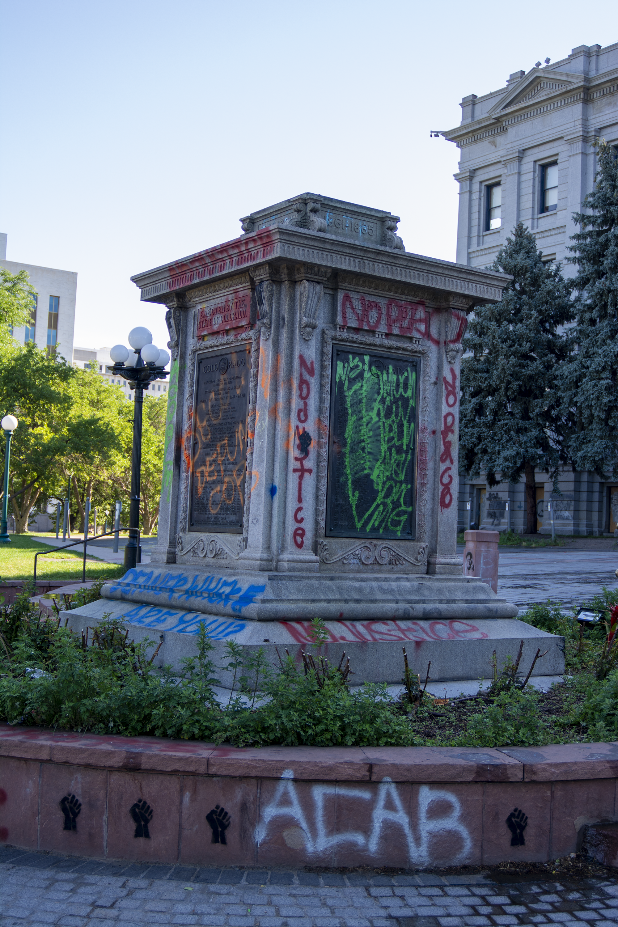 Photo of a stone pedestal that used to have a statue atop. The pedestal has been spray painted with slogans used during racial justice protests, such as "No Justice, No Peace," "ACAB," and "No Justice" is painted in several locations on the gray stone. The pedestal is surrounded by small green plants and the sticks of small shrubs.