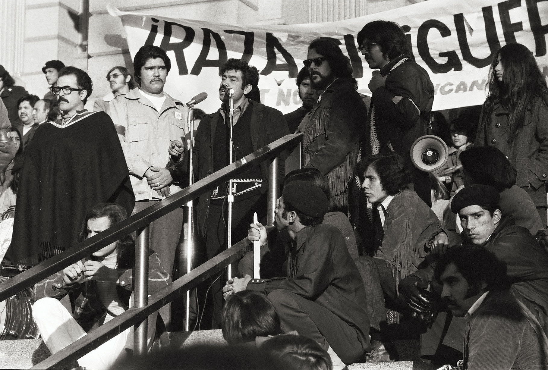 Photo of an outdoor anti-war protest rally featuring speaker Corky Gonzales. He stands in front of a microphone flanked by unidentified men and women standing and sitting on the steps. A banner behind Corky reads "RAZA SI, GUERRA NO."