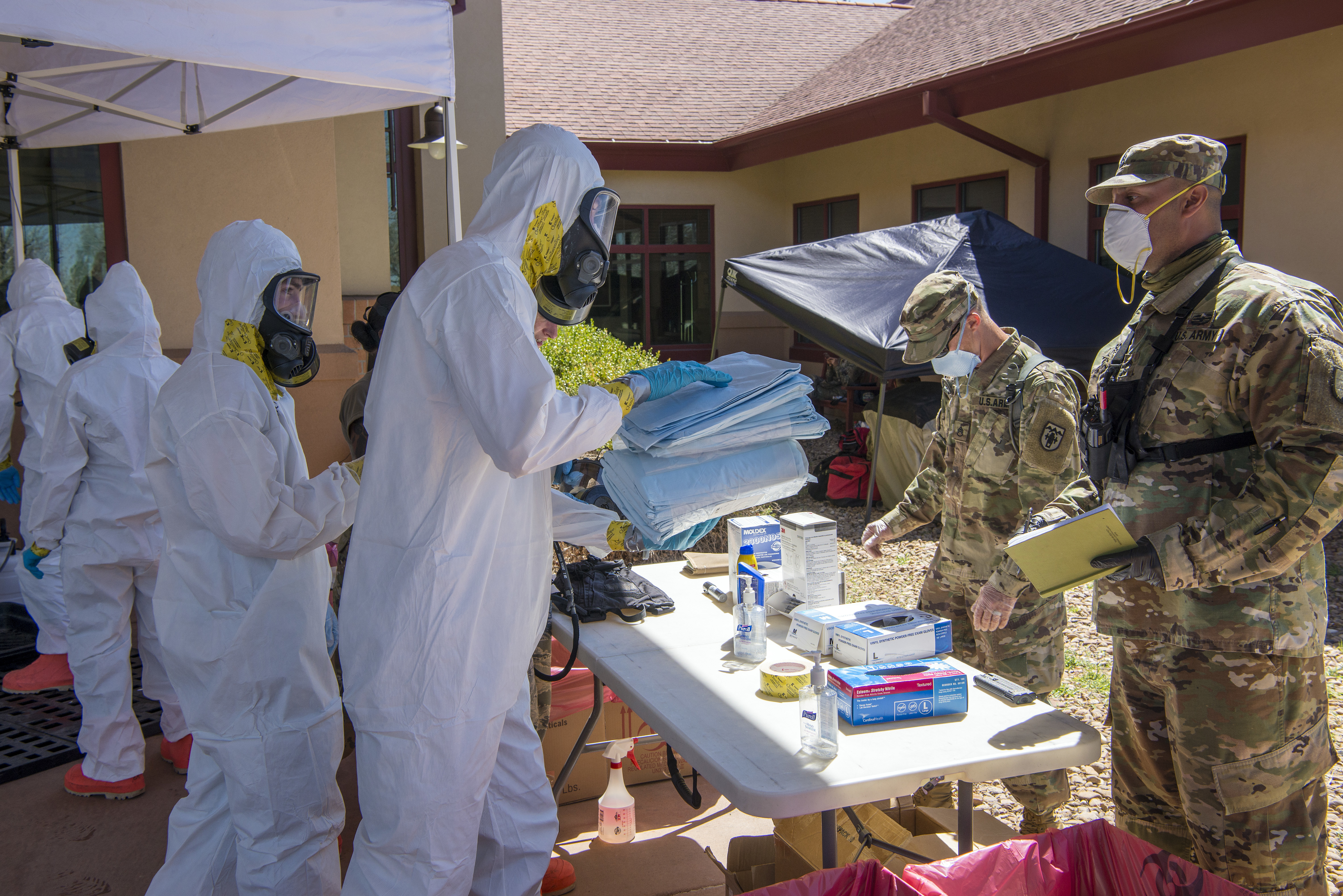 Photo of Colorado Air National Guard members, dressed in both combat fatigues and Covid-19 face masks, as well as members dressed in white decontamination suits with face masks and respirators.  They are sorting out supplies to conduct Covid-19 testing at the veterans home which is the building in the background.