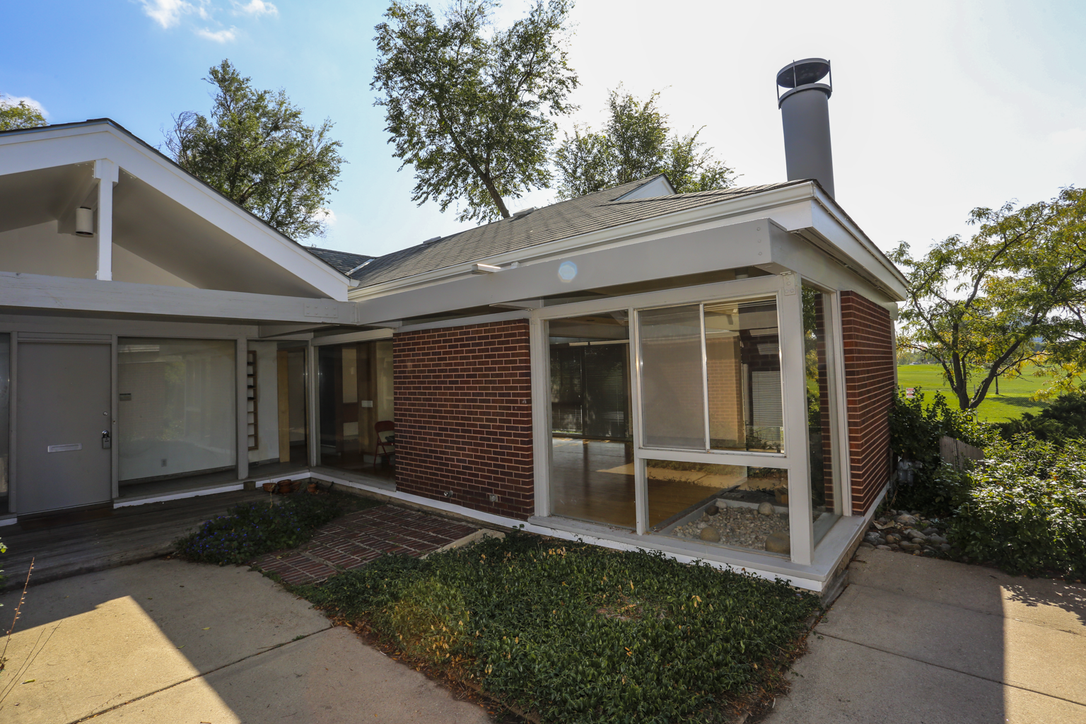 Photo of a midcentury home in Denver. The one-story building is made of red brick with white trim and windows which make up partial sections of exterior walls of the home. The windows are free of visible interior window coverings, and a few minimal pieces of furniture can be seen indoors. The home is landscaped using low-lying green shrubbery, laid brick walkways, and a large concrete pad in front of the home. The front door is large and white, with only a lock, knob, and mail slot decorate the door.