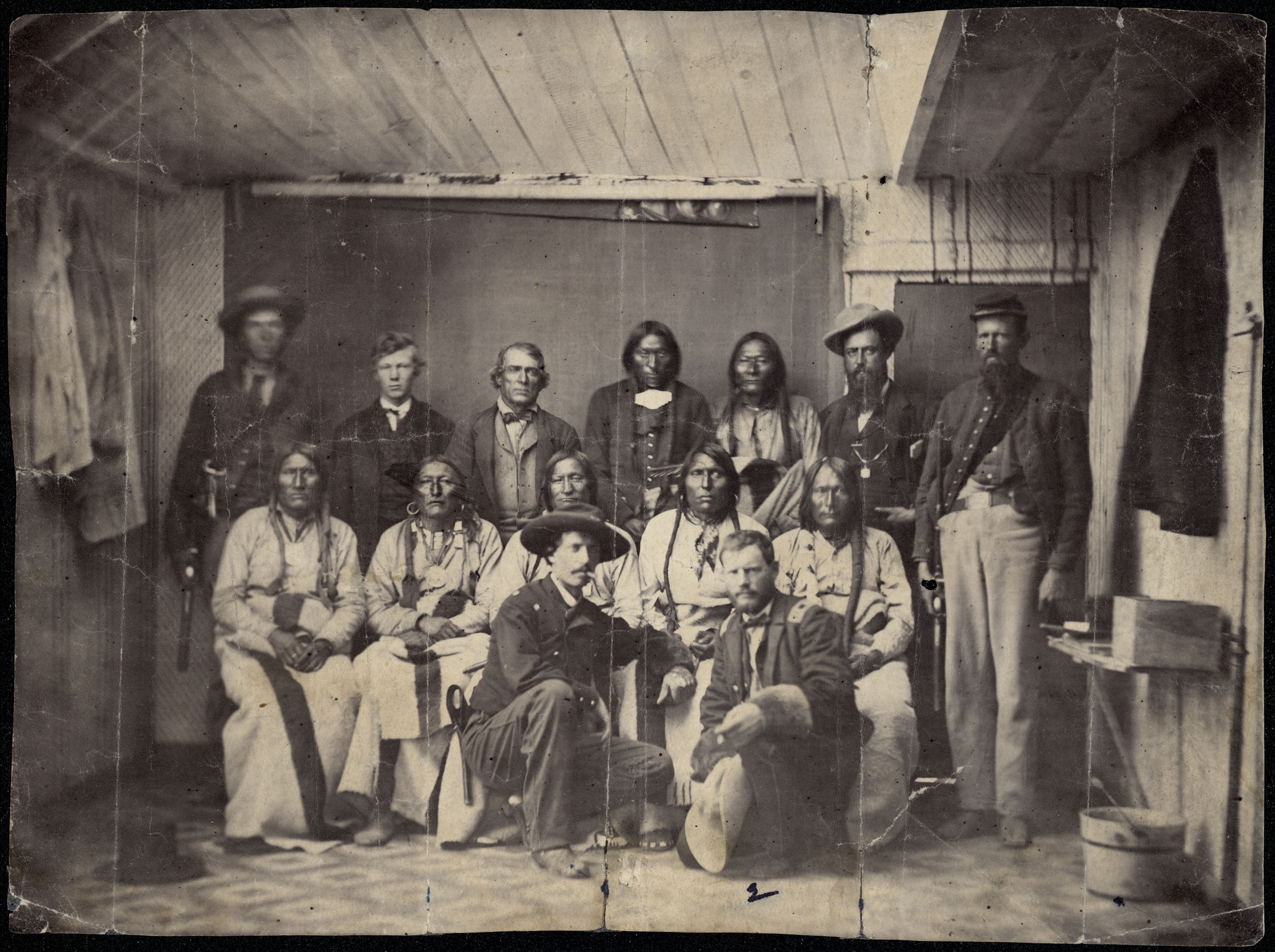 Black and white group portrait of 14 men. Two soldiers kneel in front of five seated Indigenous people. Behind them stand seven men, both Indigenous people, and soldiers.