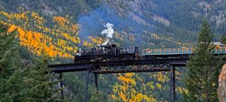 The Georgetown Loop train crosses a bridge over a lush autumn landscape of evergreens and yellow and orange aspen trees.