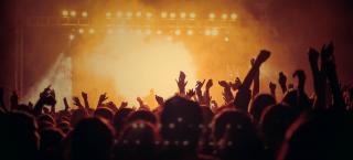 Photo of an illuminated concert stage filled with smoke, and the silouettes of concert-goers in the foreground.