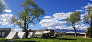 A lush green landscape with two small buildings and a few tent structures, nestled between two tall trees. A large snow capped mountain range lines the background.