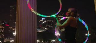 Photo of a woman in the middle of a hula hoop performance. She is using two hoops of different sizes, which are illuminated by multicolored LED lights. She is wearing a black sleeveless leotard and she is wearing a mask as a coronavirus precaution.