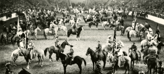 Photo of the indoor arena at the National Western Stock Show, full of dozens of riders in cowboy apparel on horseback, celebrating the inaugural event. The stands are packed with spectators.