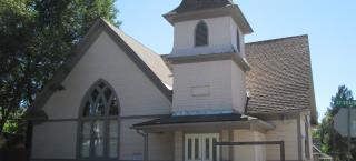 Photo of the front view of People's Methodist Episcopal Church. The building is wooden with shingled sides that are painted a tan color. The trim work is a medium brown color. There are simple wooden double doors in the front which are painted white and have twin rectangular windows. There is one large arched window to the left of the front doors,  and a small steeple directly above the  front doors. The church sits on the corner of Saint Vrain and another street, right next to the sidewalks.