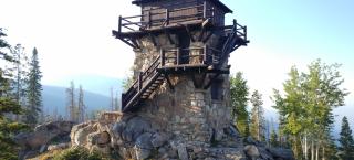 Photo of the stone and wood structure that is the Shadow Mountain Fire Lookout tower. It is a tall structure, square in shape, with a wooden deck and room that sits atop a stone base.