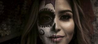 Photo of a young woman with long hair. half of her face has been painted with a skull design, with a pink flower on her forehead and gold sequins around the black circle painted around her right eye.  The right side of her mouth has vertical thin black lines across her lips. Black decorative scrolls are on her chin and right cheek.