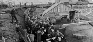 Many men seated upon the tracks in a trench along a coal mine.