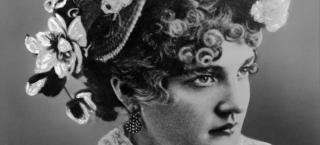 Image portrait of Baby Doe Tabor from the shoulders up. She is wearing a white lacy blouse with a flower attached on the right side of the high collar. She is wearing a very fancy hat on her head, that has white flowers on the right side of it and a large white ostrich feather on top. Her curly hair is seen beneath the hat, and she wears dangling earrings made of many small pearly beads.