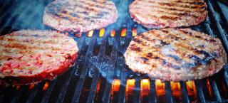 Photo closeup of four beef hamburgers cooking on a grill.  The first side of the burgers has been cooked, because the tops of each burger patty have "grill marks" lining them. The orange grill flames are visible beneath the black cast iron grates of the grill and a few wisps of smoke are rising from the cooking surface.
