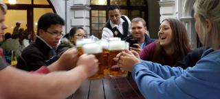 Photo of a group of friends gathered around a long wooden table at a bar or restaurant. The are each holding up a large mug of beer to the center of the table and offering a toast. There is a server at the end of the table, picking something up from the tabletop although it is obscured by the mugs of beer in the forefront. Behind the table of friends, white stone walls of the building are seen, as well as an arched doorway, and other patrons. 