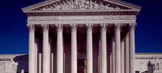 Photo of the west pediment of the Supreme Court Building in Washington DC.  The Image shows the monumental building of white marble, with its columns and massive staircase leading up to the door. Seated statues are seen toward the top of either side of the staircase. Above the columns, the pediment features a sculptural group of nine figures along with the words, "Equal Justice Under Law."