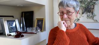 Photo of Jean Dubofsky, sitting in her office. The walls and shelves behind her are white, contrasting her orange sweater and red-framed eyeglasses. There are gold and silver-framed photos on the shelves, and a painting on the wall behind her. She is resting her chin on her closed right hand, and she is looking straight ahead at the camera.