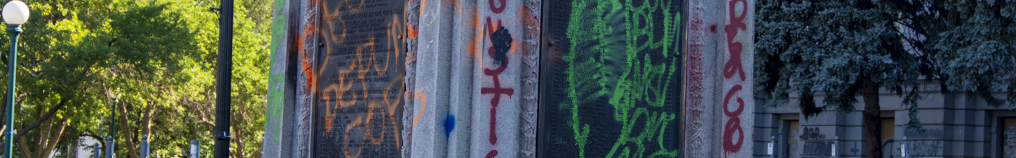 Photo of a stone pedestal that used to have a statue atop. The pedestal has been spray painted with slogans used during racial justice protests, such as "No Justice, No Peace," "ACAB," and "No Justice" is painted in several locations on the gray stone. The pedestal is surrounded by small green plants and the sticks of small shrubs.