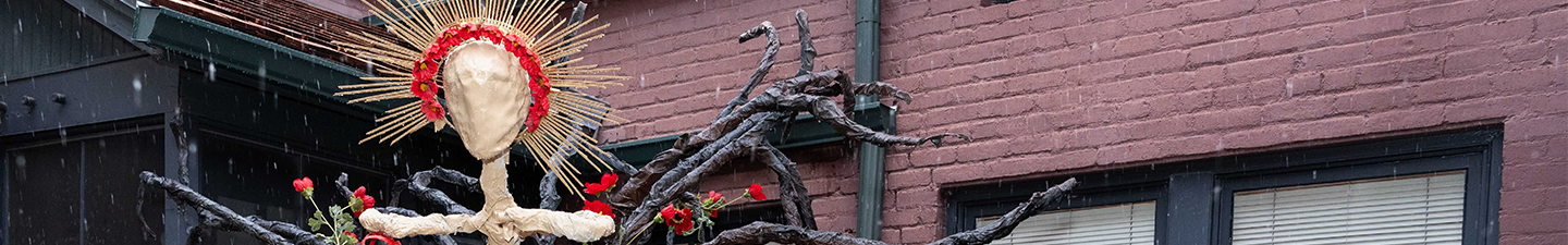 Artist Ariana “Nani” Gonzales next to her sculpture, Genesis. It shows a paper mache skeletal figure with a golden halo covered red roses, and dark branches.