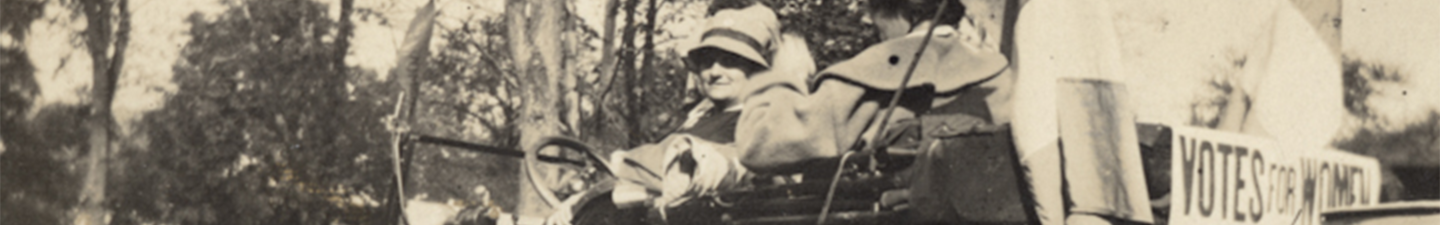 Suffragettes hang flags with "Votes for Women" on the back of an old automobile.