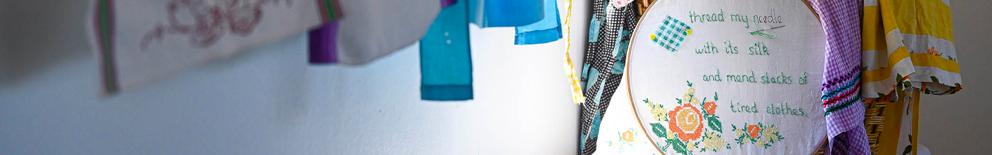 Drapes of colorful fabric fill the foreground as natural light focuses on an embroidery loop filled with flowers and the words: Thread my needle with its silk and men stacks of tired clothes.