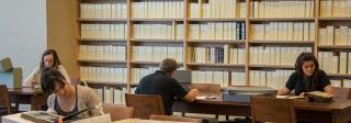Four people sitting at desks, looking to various texts. A wall of books and periodicals on bookshelves sits behind them. 