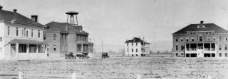 Photo of four buildings sitting on a barren piece of land. in the foreground, part of a dirt road can be seen. There are 3 vehicles parked in front of two of the buildings on the left side of the image. In the far distance, the hazy silouette of mountains can be seen. There are no people immediately visible in this historic photograph.