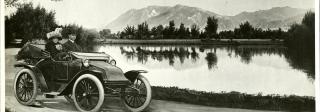 A man and a woman in turn-of-the-century dress ride in an early open-top car. The car is driving along a gravel path next to a small body of water. Mountains are on the horizon. A caption reads "The 100-mile Fritchle: The recognized highest-grade electric automobile in the world. Address: Fritchle, Denver."