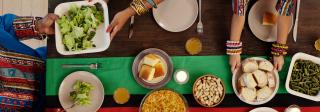 Photo looking down upon a long dining table made of dark brown wood and decorated with a long table runner down the center consisting of wide green and red stripes divided by a thin black stripe of fabric. Three people in colorful attire sit around the table, passing dishes of food and serving some onto plates. Dishes of greens, cornbread, green beans, sliced bread, and other foods are being shared.