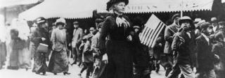 A woman in a black dress walks down a street. Children and women march with her carrying picket signs.