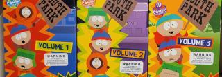 A photograph taken with a cell phone camera, of three VHS tapes in their cardboard covers, lined up on white metal shelving so that the full cover of each tape is visible. The tapes are recordings of the "South Park" animated television series, Volumes 1, 2, and 3. Each cover prominently displays the "TV-MA" warning in the upper right corner, as well as a warning box below the volume number, indicating that these programs are for mature, audiences only and contain adult language and situations.