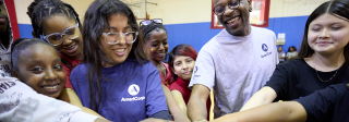 A group of children and 2 AmeriCorps members stand in a gym smiling. They are huddled in a circle putting their hands together.
