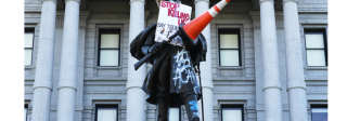 Image of a vandalized statue outside of a government building. Signs and graffiti say "Stop killing us" and "ACAB"