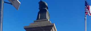 Profile view of a dark stone statue depicting the bust of Christopher Columbus. The American flag waives behind it.
