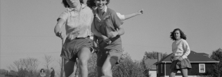 Black and white image of three young women kicking a ball in a grass field. There's houses and two adults behind them.