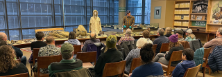 A large group of visitors in a rows of chairs sit attentively while a presenter shares numerous artifacts, including many uniforms from the 10th Mountain Division.