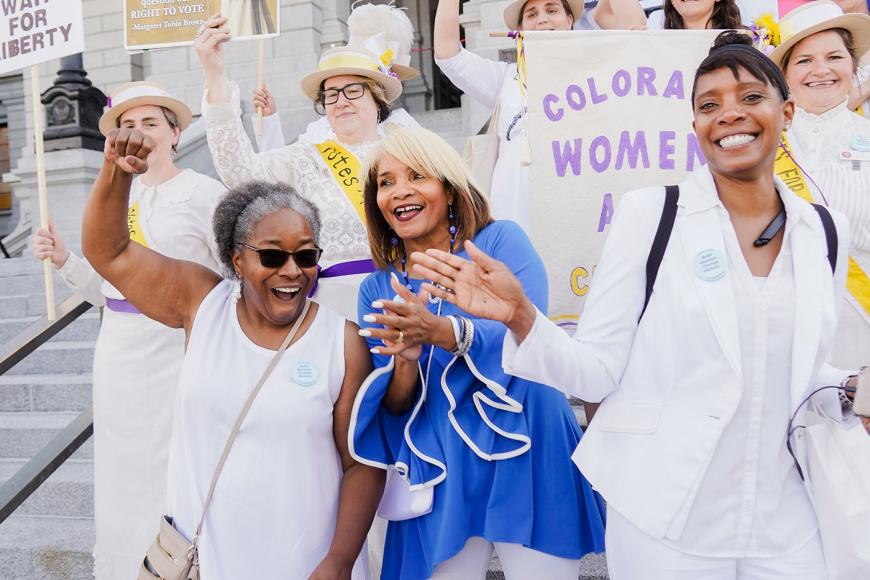 A large group of women gathered on the steps of the Capitol with protest signs and smiling.