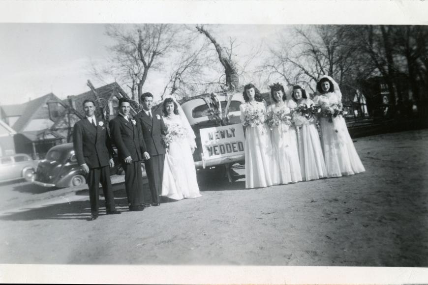 A black and white photo of a wedding party gathered at a car with the sign "Newly Wedded" on the back.