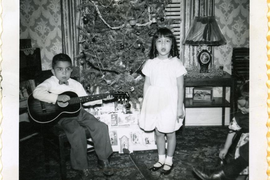 Two children standing in front of a Christmas tree. The boy is playing a guitar and the girl is singing.