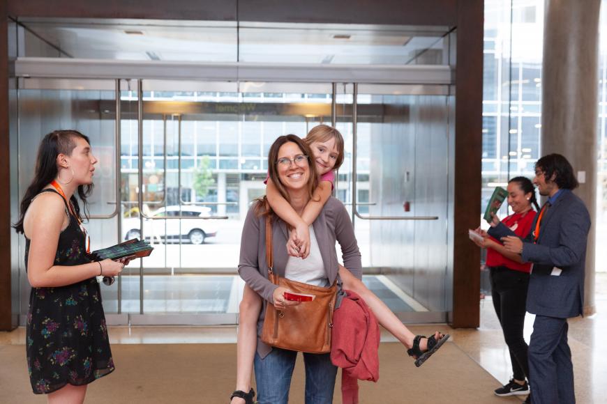 A History Colorado member and her daughter enter the History Colorado Center, smiling. The child is riding on her mother's back.