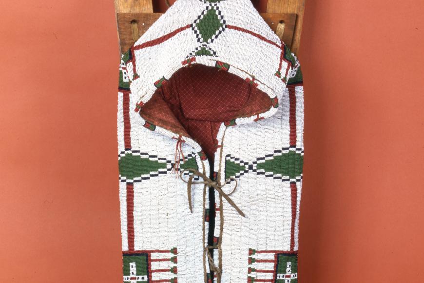 Decorative handcrafted cradleboard. White, red and green stitch textiles, mounted on two long adorned pieces of wood.