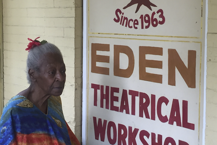An elderly Black woman, Lucy Walker, is seated in a colorful shawl before a sign which reads Eden Theatrical Workshop (since 1963)