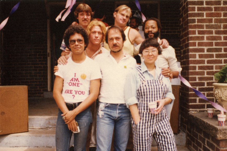 A group of seven adults gathered on the steps of a brick building. One person in front wears a shirt that reads: "I am one. Are you?"