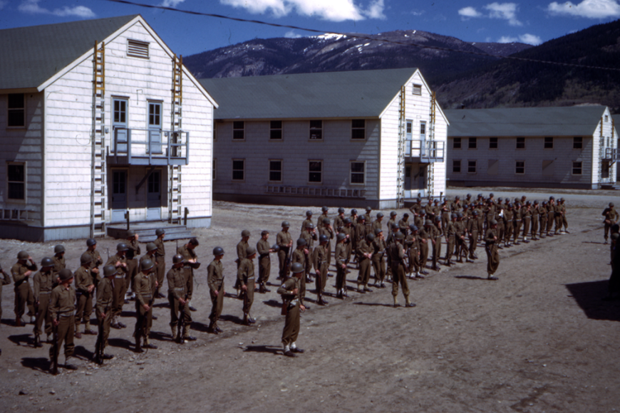 A large group of soldiers in uniform at Camp Hale, standing in formation in front of three large, white barracks.